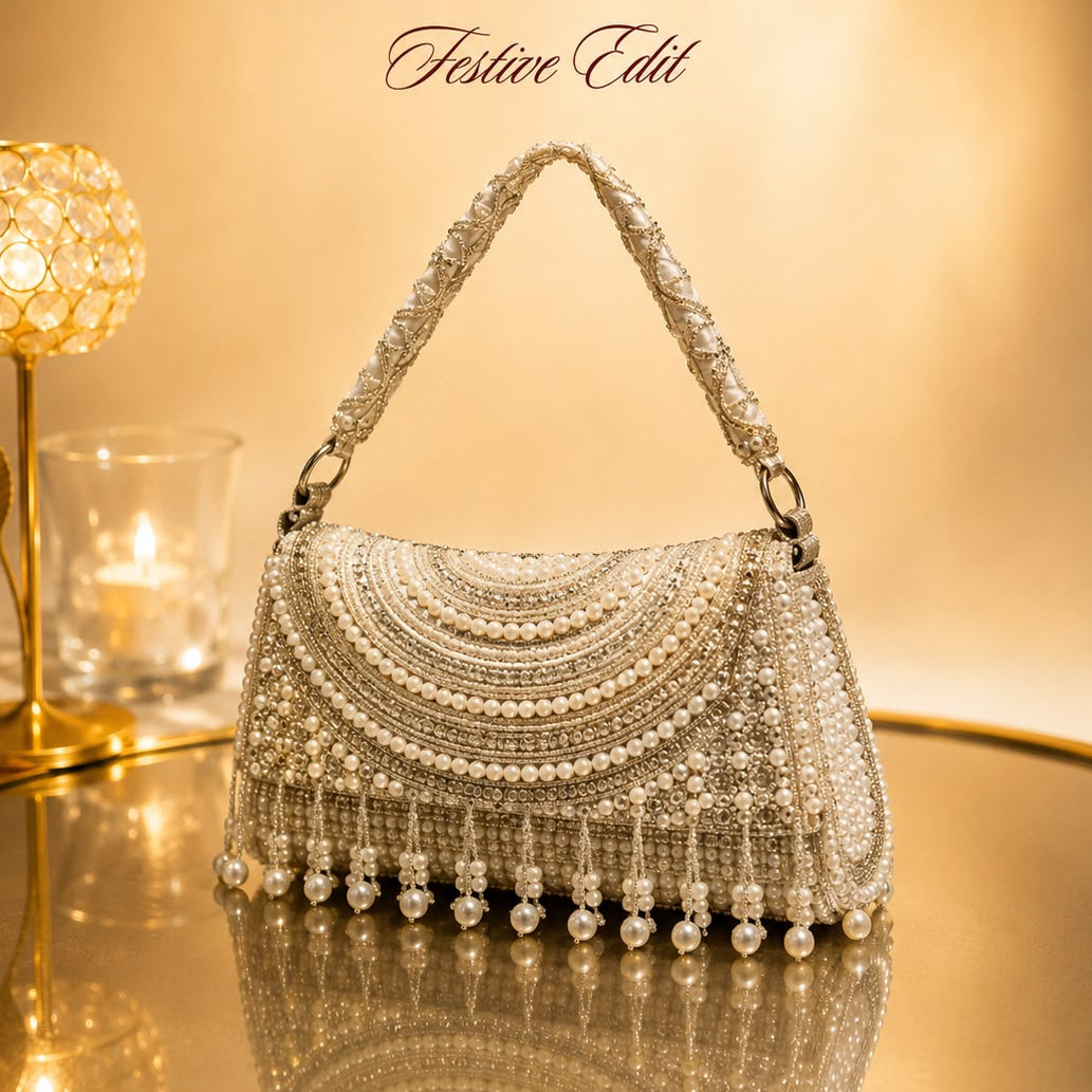A heavily embellished silver and white beaded handbag with a pearl fringe, displayed on a reflective gold surface next to a lit decorative lamp. The brand logo 'The Nukkad Store' is visible at the top.