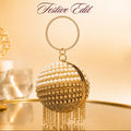 A gold and pearl beaded spherical clutch purse with hanging fringe, displayed on a reflective surface with a warm glowing background. The Nukkad.